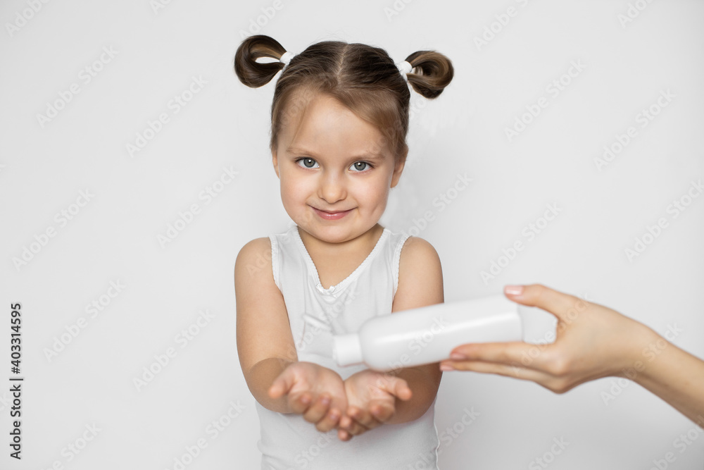 A little three years old girl with blonde hair, standing on isolated white background, looking at camera with smile, while hand of her mother pouring cream milk or body oil on her hands A little three years old girl with blonde hair, standing on isolated white background, looking at camera with smile, while hand of her mother pouring cream milk or body oil on her hands