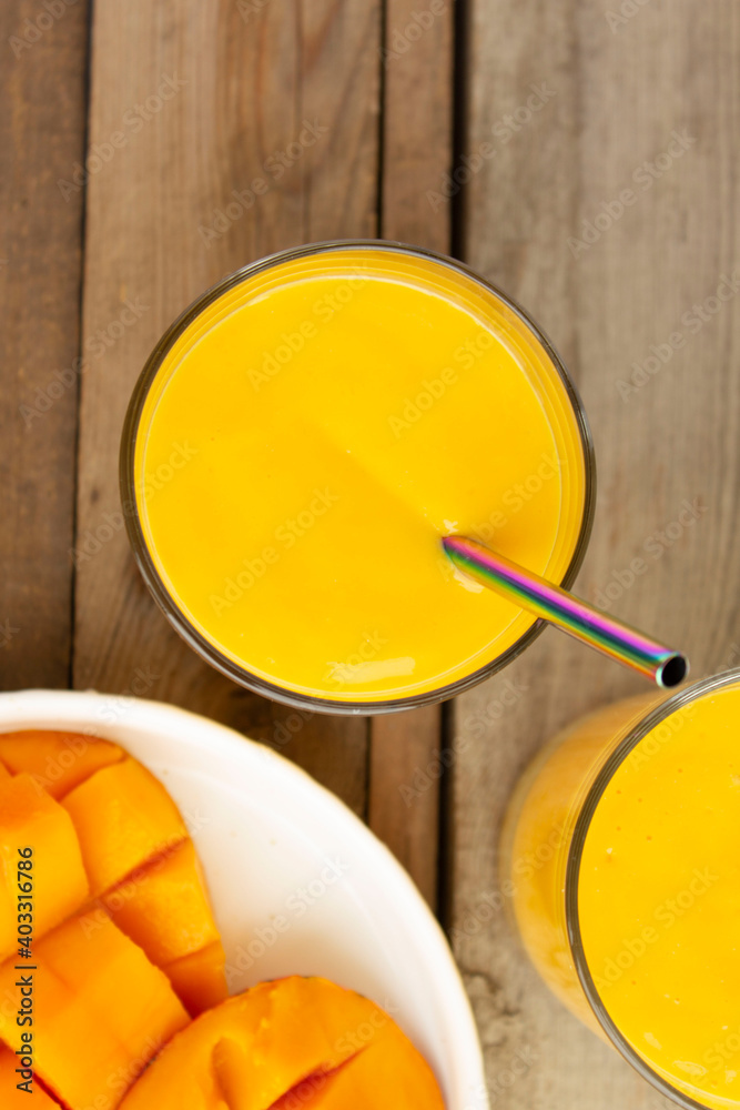 Mango lassi top view. Mango drink in the glass with multicolored metal or eco straw. White plate with sliced mango fruit on wooden table Mango lassi top view. Mango drink in the glass with multicolored metal or eco straw. White plate with sliced mango fruit on wooden table