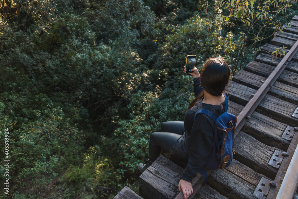 Adventurous girl taking a picture with her cell phone of the landscape at sunset in the forest, sitting on a bridge of the train tracks (POV photo) Adventurous girl taking a picture with her cell phone of the landscape at sunset in the forest, sitting on a bridge of the train tracks (POV photo)