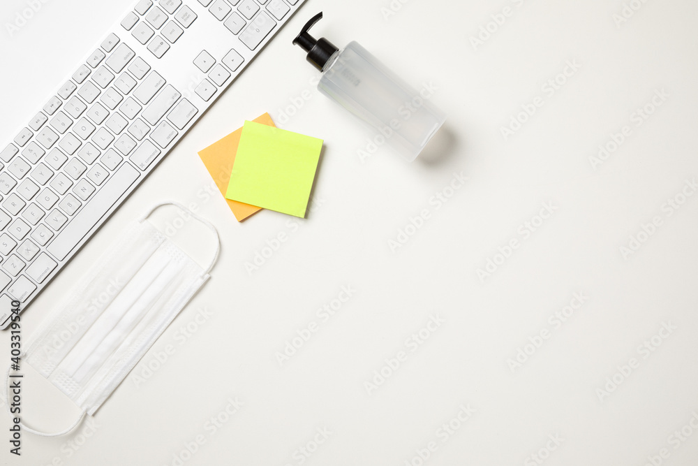 Top view of a white table with a keyboard, a white surgical mask, two colored reminder stickers and a alcohol gel pump dispenser. With copy space. Top view of a white table with a keyboard, a white surgical mask, two colored reminder stickers and a alcohol gel pump dispenser. With copy space.