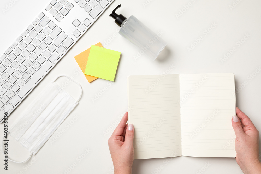 Obraz premium Top view of a white table with a keyboard, a white surgical mask, two colored reminder stickers, a alcohol gel pump dispenser and female hands holding a blank notebook. With copy space.