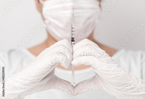 Close up of doctor or nurse wearing white gloves, making a heart shape with hands, holding a syringe with a drop in the tip of the needle, in white background.