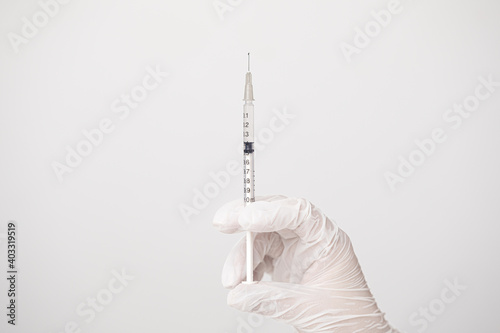 Close up of doctor or nurse hand wearing white gloves, holding a syringe with a drop in the tip of the needle, in white background.