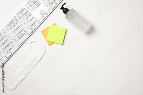 Top view of a white table with a keyboard, a white surgical mask, two colored reminder stickers and a alcohol gel pump dispenser. With copy space.