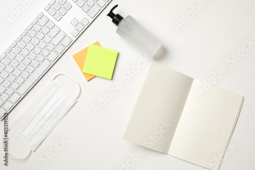 Top view of a white table with a keyboard, a white surgical mask, two colored reminder stickers, a alcohol gel pump dispenser and a open blank notebook. With copy space.
