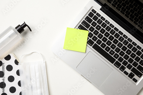 Top view of white table workspace with a laptop, a yellow reminder sticker, a white surgical mask, alcohol gel pump dispenser and a dotted notebook. Copy space.