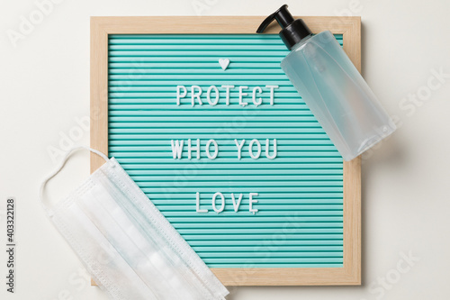 Top view of a frame with protect who you love written, a white surgical mask and a alcohol gel pump dispenser.