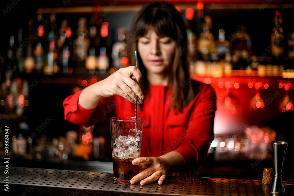 transparent glass with drink stands on bar and woman bartender is stirring it with spoon