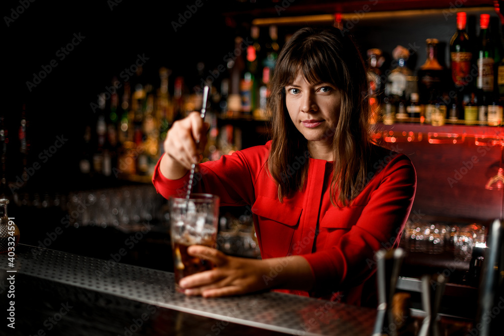 woman bartender stands behind bar and mixes cocktail in glass with spoon