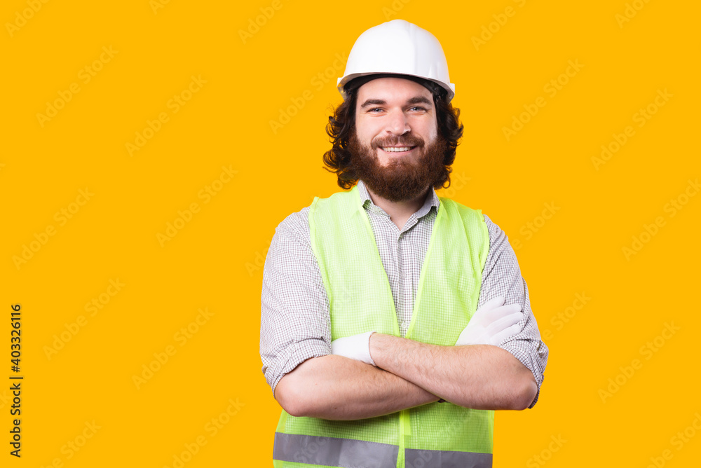 A awesome portrait of a young bearded engineer looking at the camera with his arms crossed near a yellow wall .