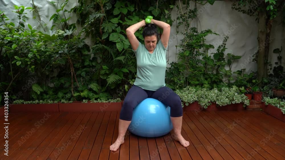 Mujer haciendo ejercicio con mancuernas sentada en una pelota  en el jardín de su casa
