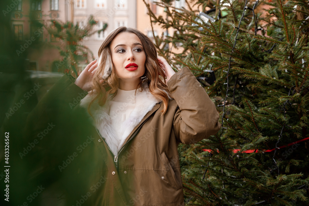 Lady in a knitted sweater and fluffy headphones. Outdoor portrait of young beautiful happy smiling girl is wearing fur coat near decorated Christmas tree. Lady in a knitted sweater and fluffy headphones. Outdoor portrait of young beautiful happy smiling girl is wearing fur coat near decorated Christmas tree.