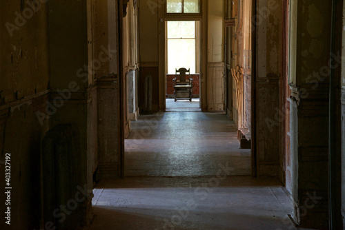 darkened hallway without electricity  light coming in through a dirty window at the end of the hallway  one chair sitting empty under the window.