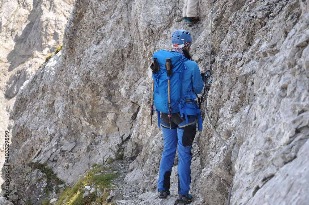 Kobieta ubrana na niebiesko idzie wąską, skalną półką na via ferracie w Dolomitach