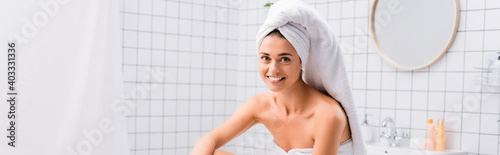 happy young woman with white towel on head smiling at camera in bathroom, banner