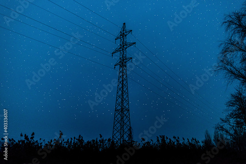 silhouette of high voltage electric power transmission lines tower, in the middle of a field against a blue night sky with stars
