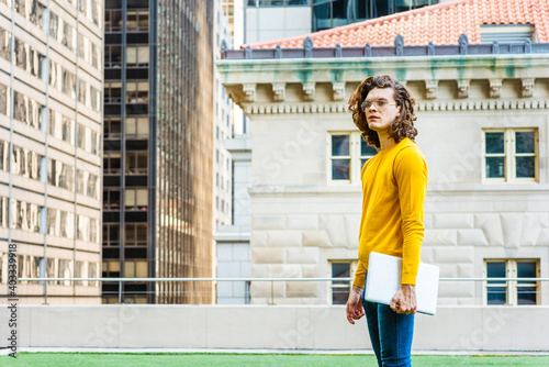 Photography Hispanic American College Student Studying in New York City, with brown curly hair, wearing glasses, yellow long sleeve T shirt, jeans, holding laptop computer, standing on green lawn, thinking