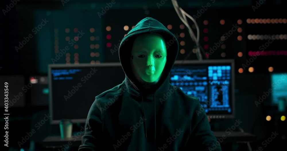 Close-up Shot of a Masked Hacker in a Hoodie Sitting in the Middle of Data Center full of Rack Servers. Close-up Shot of a Masked Hacker in a Hoodie Sitting in the Middle of Data Center full of Rack Servers.