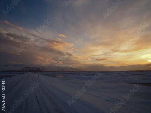 Iceland Ring Road (Route 1) covered in snow at sunset, lighting clouds in orange and golden colours, driving to the mountains in the distance