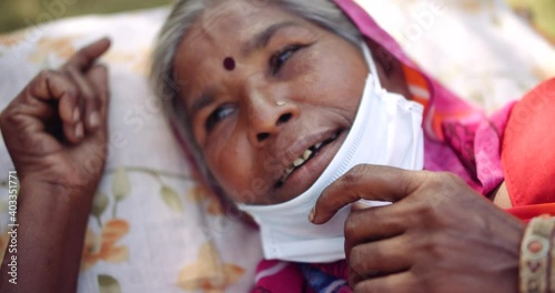 Slow-motion portrait top-shot of a senior woman smiling positive emotion outdoors wearing surgical faec mask in pain disease illness on her bed coughs sick being attended by healthcare workers  photo