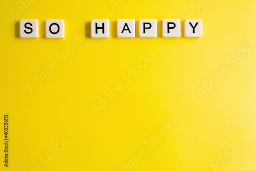 the words so happy written as a flat lay in wood scrabble tiles on a plain yellow background