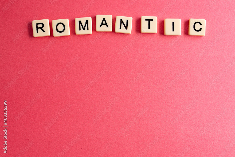 the word romantic written as a flat lay in wood scrabble tiles on a plain red background surrounded by red, white, and pink candies the word romantic written as a flat lay in wood scrabble tiles on a plain red background surrounded by red, white, and pink candies