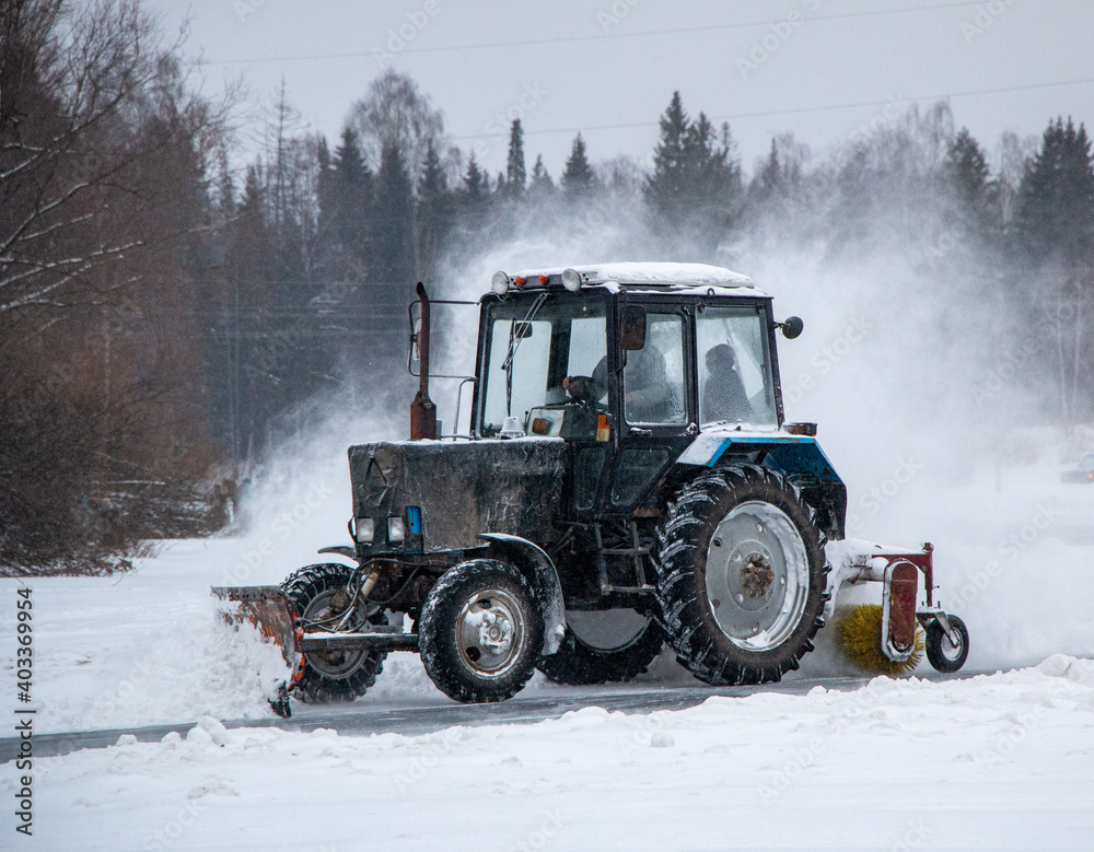 an old snow-covered tractor with blade removes snow from the road, sweeps snow from the road with large brush, in severe blizzard. an old snow-covered tractor with blade removes snow from the road, sweeps snow from the road with large brush, in severe blizzard.