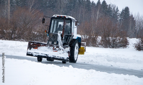 an old snow-covered tractor with blade removes snow from the road, sweeps snow from the road with large brush, in severe blizzard.