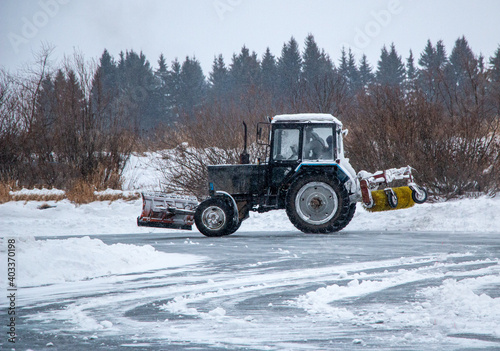 an old snow-covered tractor with blade removes snow from the road, sweeps snow from the road with large brush, in severe blizzard.