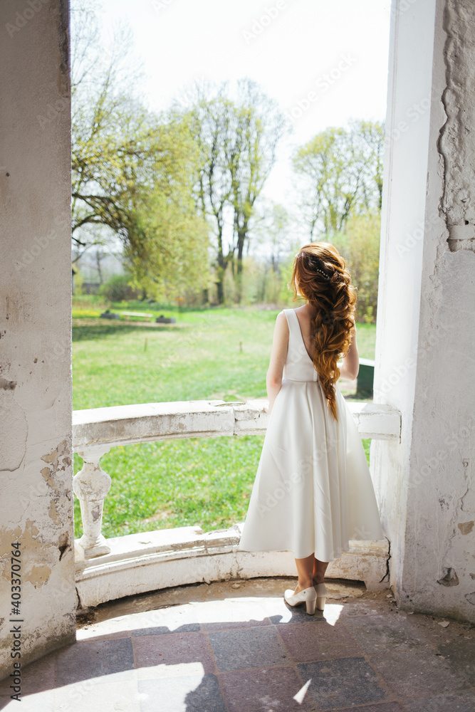 Fototapeta premium A woman with long red hair. A woman in a white dress stands on the porch. Beautiful hairstyle of long hair.
