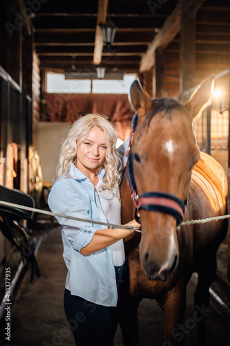 A pretty blonde dressed in a shirt stands in the stable next to a horse.