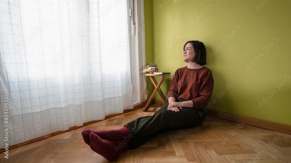 Smiling woman sitting on the wooden floor of her room looks at the window with white curtains and a wooden bench with a green notebook and a cactus Smiling woman sitting on the wooden floor of her room looks at the window with white curtains and a wooden bench with a green notebook and a cactus