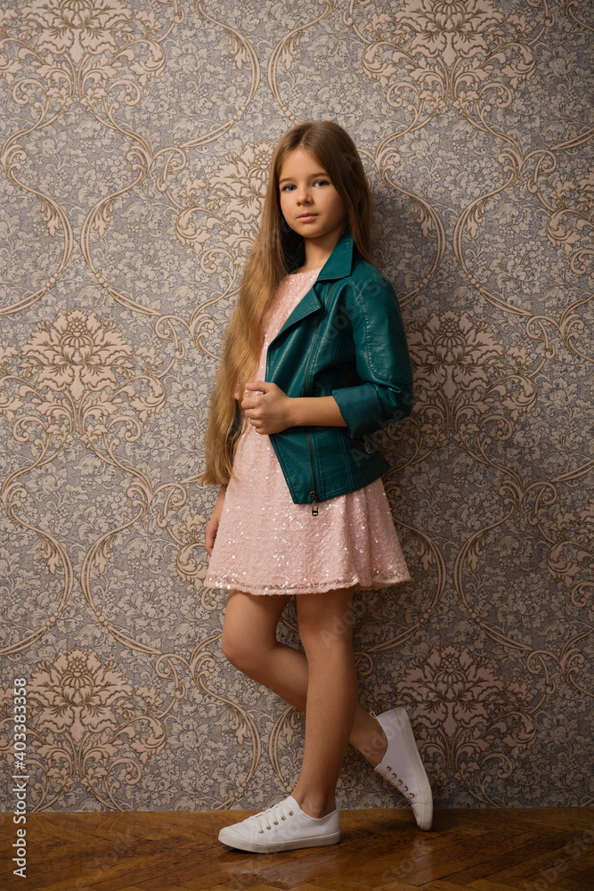 beautiful child girl with long beautiful hair in a pink dress and a bright leather jacket against the background of a wall with wallpaper