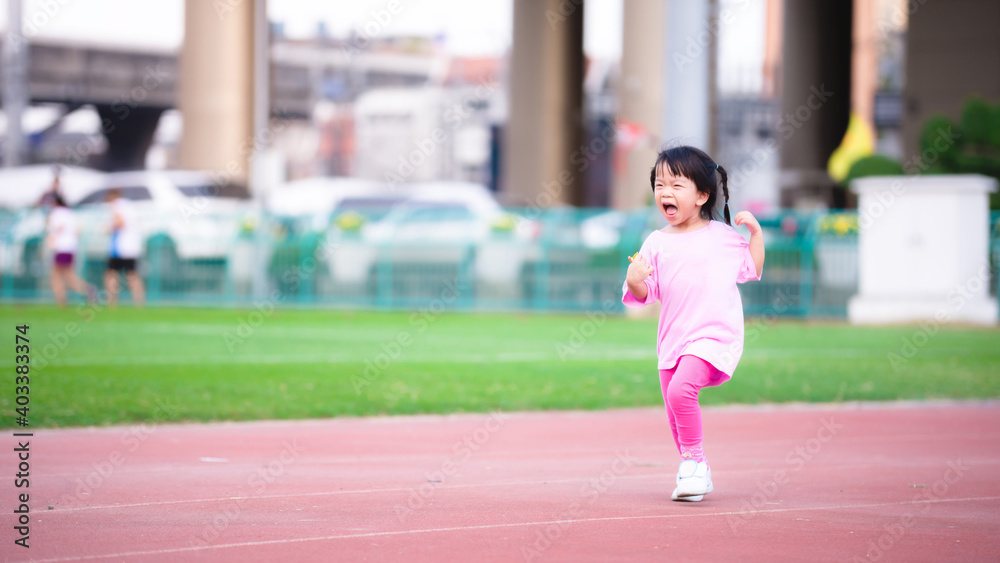 The agile girl was running happily on the track of the stadium. Happy child is happy to exercise. Cute kid wearing a pink dress and white sneakers. Active children are 3 years old.