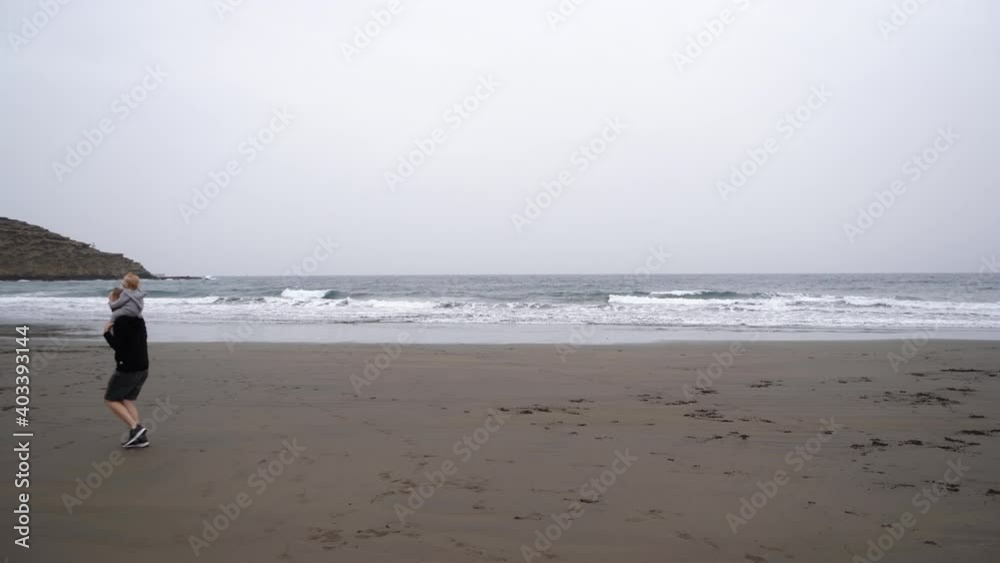 Father runs with a small child around his neck on a sandy beach by the ocean. Cloudy windy cold weather and a family walk. Happy dad and daughter. Father runs with a small child around his neck on a sandy beach by the ocean. Cloudy windy cold weather and a family walk. Happy dad and daughter.
