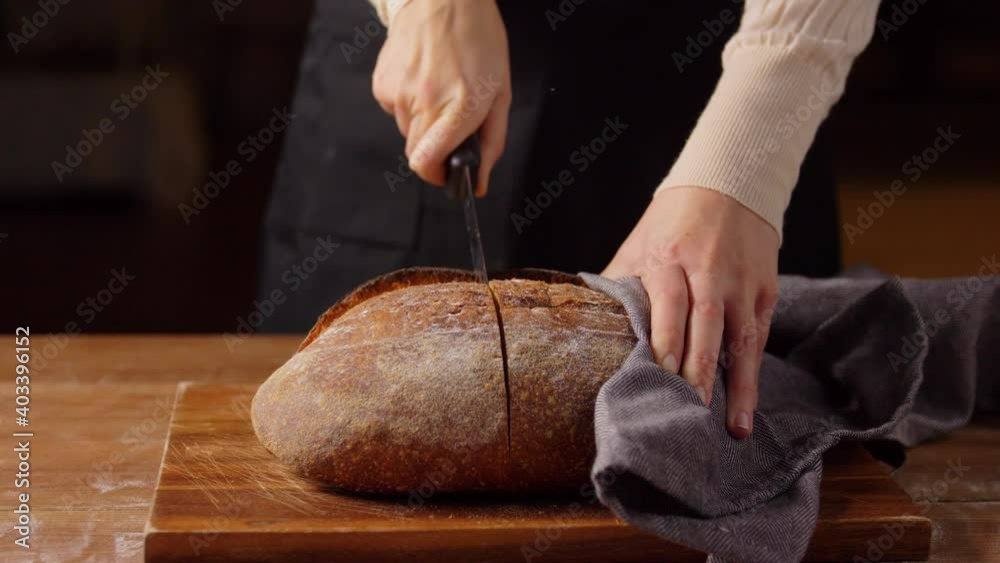 food cooking, baking and people concept - female baker with knife cutting loaf of bread into halves at bakery or kitchen