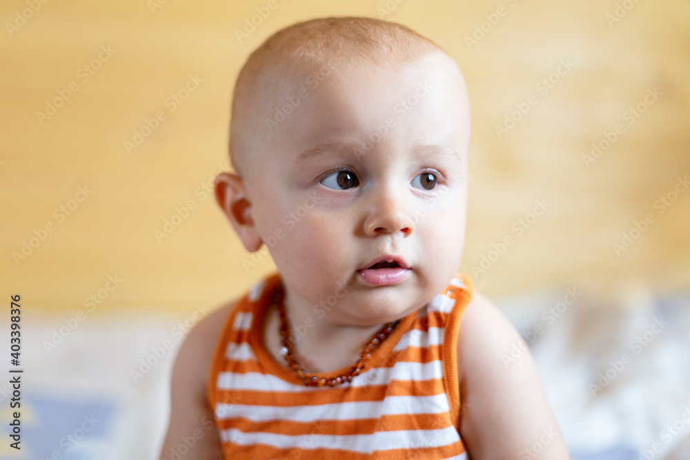 A portrait of a cute adorable curious baby boy of 12 months or 1 year old looking aside wearing amber teething baby necklace in a sleeveless striped orange top. A portrait of a cute adorable curious baby boy of 12 months or 1 year old looking aside wearing amber teething baby necklace in a sleeveless striped orange top.