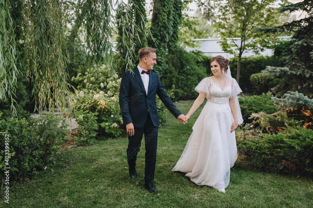A young groom in a blue suit walks through the park with green plants, holding the hand of a beautiful curly-haired bride in a long white dress. Wedding portrait of newlyweds in love. A young groom in a blue suit walks through the park with green plants, holding the hand of a beautiful curly-haired bride in a long white dress. Wedding portrait of newlyweds in love.
