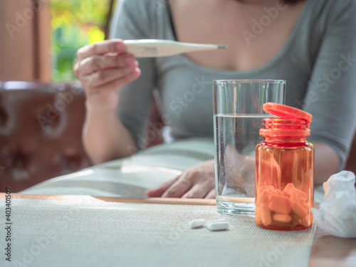 virus protection concept, glass of water, orange bottle of pills in blurry background of a woman looking at thermometer, protect for coronavirus or flu.