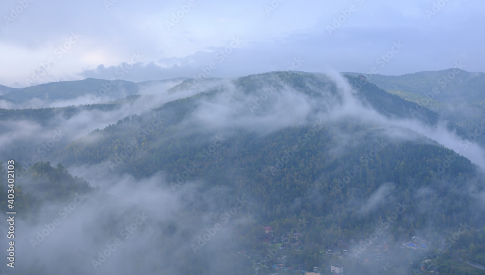 Fog in the mountains. Green forest with golden colors. Residential buildings in the valley.
