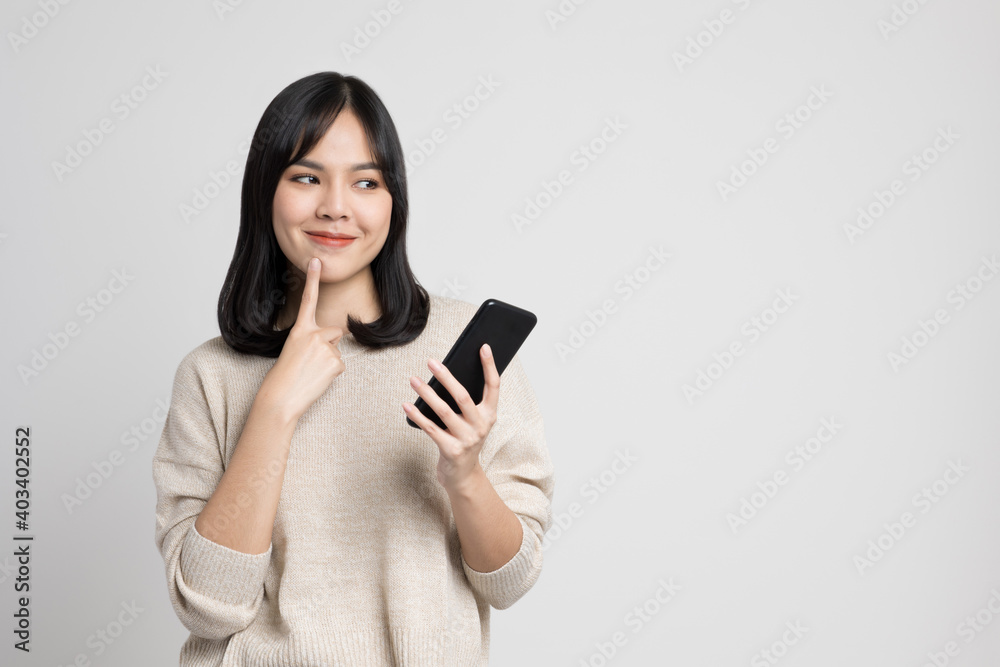 Beautiful young asian woman holding smartphone standing on isolated white background. She is looking at blank space and thinking something. Beautiful young asian woman holding smartphone standing on isolated white background. She is looking at blank space and thinking something.