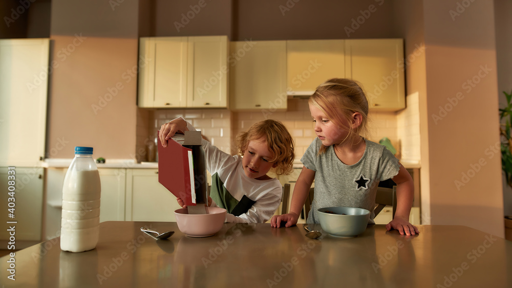 Joyful little boy pouring chocolate flakes into a bowl while preparing cereal with milk for breakfast, sitting together with his sister at the table in the kitchen Joyful little boy pouring chocolate flakes into a bowl while preparing cereal with milk for breakfast, sitting together with his sister at the table in the kitchen