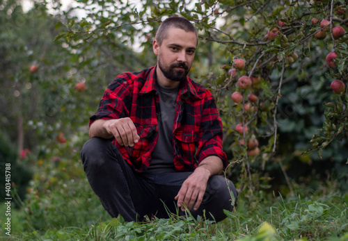 Portrait of a sitting bearded gardener against the background of trees with apples. Organic farm concept. Close-up © Serhii