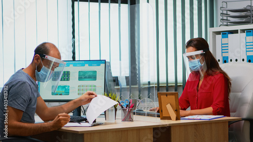 Employees wearing mask and visor sitting in new normal office room respecting social distancing. Coworkers working in modern workplace respecting protection rules against covid virus using plexiglass.