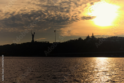 Majestic sunset landscape. Silhouettes of famous Kyiv's hills with Mothercare Monument and church from Kyiv Pechersk Lavra. Dramatic sky with vibrant clouds and bright sun. Bare trees on the hill © evgenij84