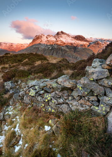 Old rural stone wall leading to view of snowcapped Cumbrian mountains; The Langdale Pikes as morning sunlight casts a pink glow. Lake District, UK.