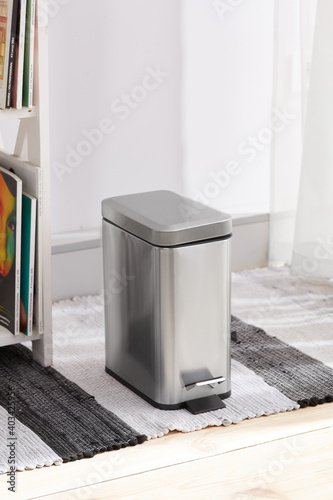Subject shot of lidded hands-free wastebasket with foot pedal. The stainless steel trash can is located on striped white and gray carpet near bookshelf in the light room.