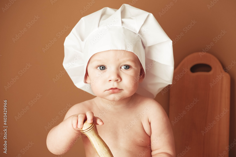 Portrait little baby boy in a chef's hat sitting on the table holds a wooden rolling pin on a brown background. Copy, empty space for text Portrait little baby boy in a chef's hat sitting on the table holds a wooden rolling pin on a brown background. Copy, empty space for text