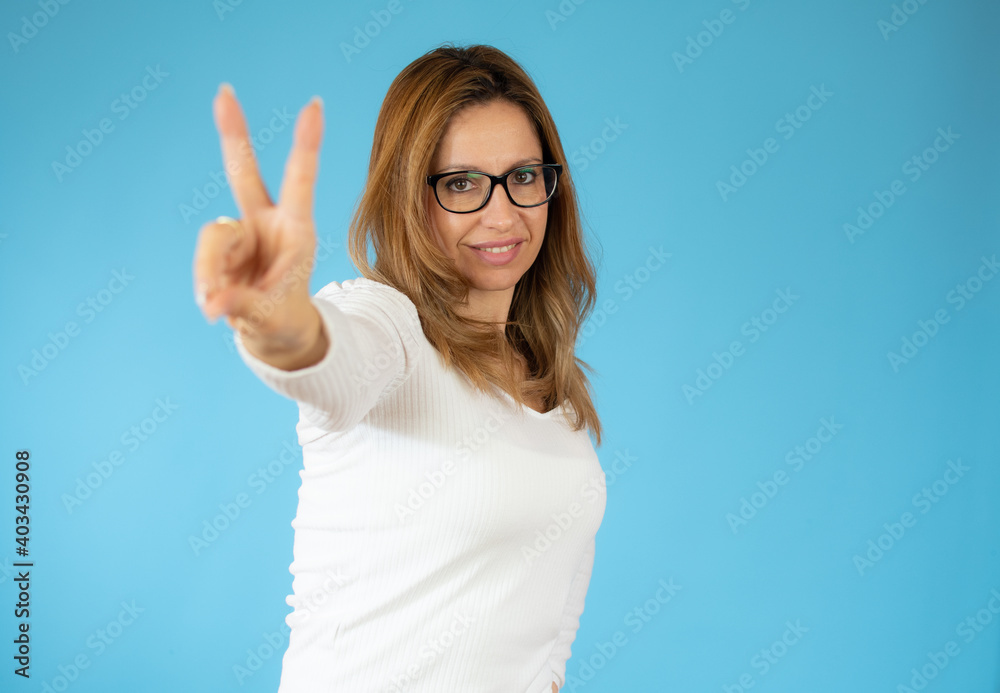 Portrait of gorgeous woman looking at camera with smile and showing peace sign with fingers isolated over blue background Portrait of gorgeous woman looking at camera with smile and showing peace sign with fingers isolated over blue background