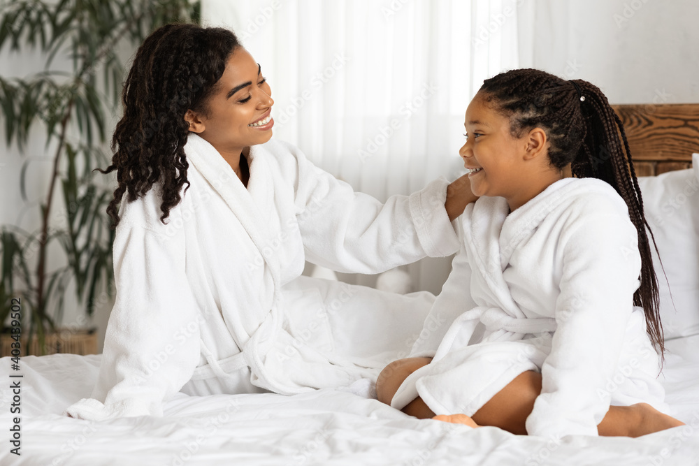 Cheerful African American Mom And Daughter In Bathrobes Sitting On Bed, Relaxing Cheerful African American Mom And Daughter In Bathrobes Sitting On Bed, Relaxing